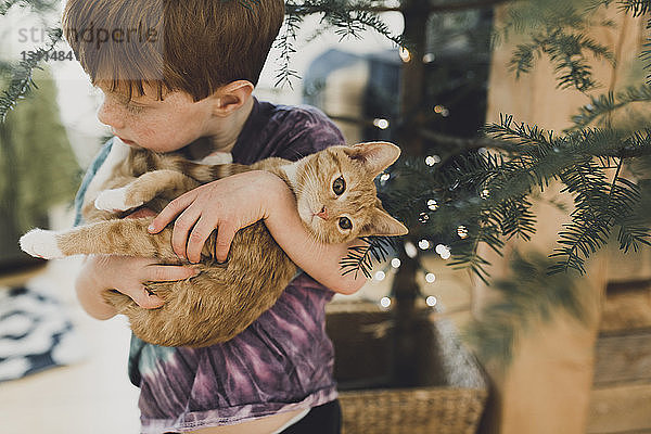 Schrägansicht eines Jungen  der ein süßes Kätzchen trägt  während er zu Hause an der Pflanze steht
