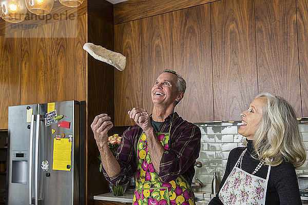 Fröhlicher Mann wirft Pizzateig  während eine Frau in der heimischen Küche das Essen zubereitet