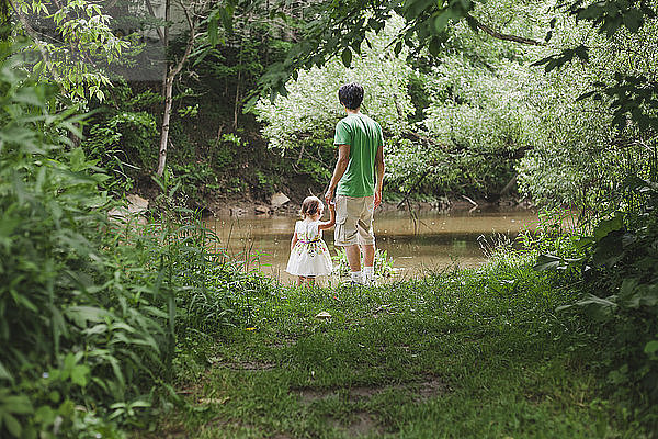 Rückansicht eines Vaters mit Tochter auf einem Grasfeld am Fluss im Park