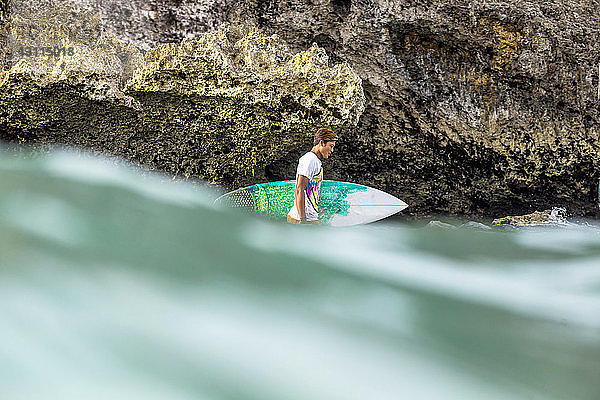 Seitenansicht eines Mannes mit Surfbrett  der an einer Felsformation im Meer steht