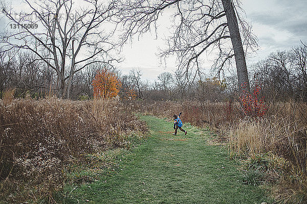 Glücklicher Junge mit ausgestreckten Armen rennt auf Grasfeld gegen den Himmel im Wald
