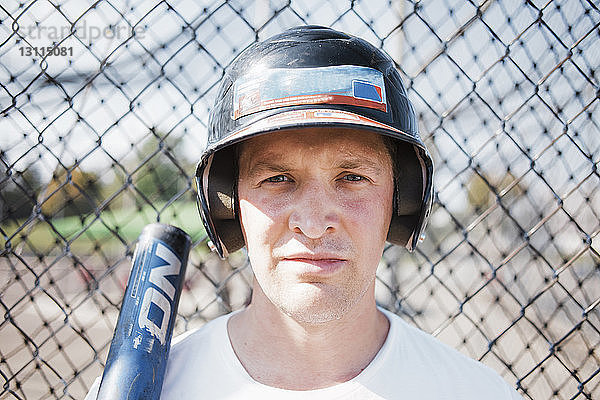 Nahaufnahme eines selbstbewussten Baseballspielers mit Sporthelm gegen den Maschendrahtzaun auf dem Spielfeld bei sonnigem Wetter