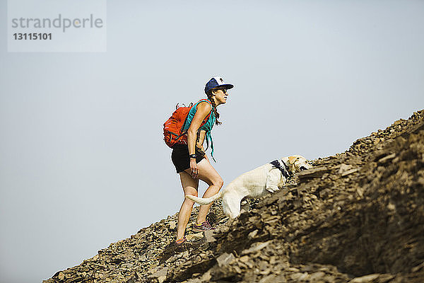 Seitenansicht einer Frau  die einen Rucksack mit Hund trägt und gegen den klaren Himmel auf einen Berg klettert