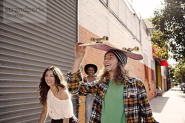Mann trägt Skateboard auf dem Kopf  während er mit Freundinnen geht