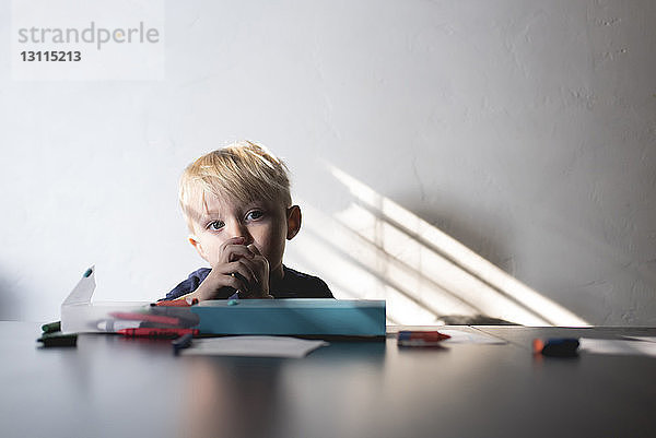 Süßer Junge mit Buntstiften schaut weg  während er am Tisch an der Wand sitzt