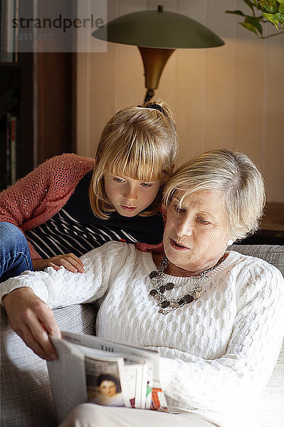 Großmutter zeigt Enkelin Zeitschrift  während sie zu Hause auf dem Sofa sitzt