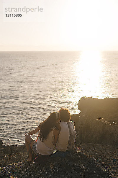 Rückansicht einer Freundin  die ihren Freund küsst  während sie bei Sonnenuntergang auf einem Felsen am Strand gegen den Himmel sitzt