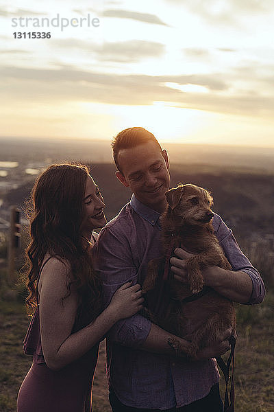 Junges Paar mit Hund steht bei Sonnenuntergang auf dem Feld