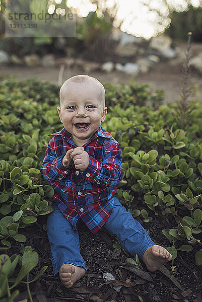 Porträt eines fröhlichen kleinen Jungen  der inmitten von Pflanzen im Park sitzt