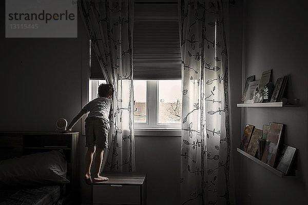 Rückansicht eines Jungen  der durch ein Fenster schaut  während er zu Hause auf einem Schrank steht