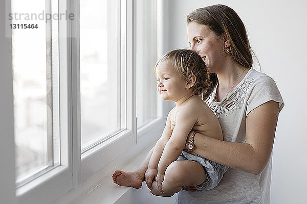 Seitenansicht einer Mutter  die eine Tochter trägt  während sie zu Hause durch ein Fenster schaut
