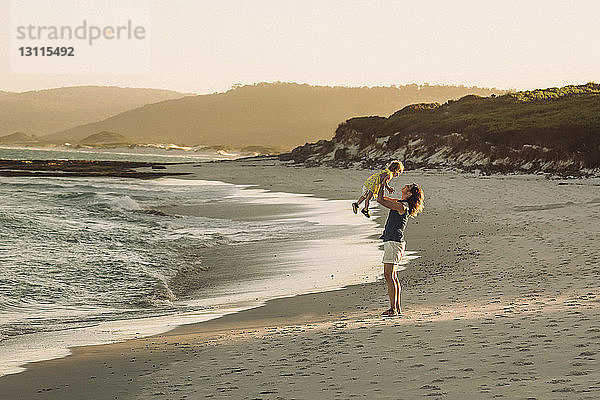 Seitenansicht einer Mutter  die eine Tochter trägt  während sie bei Sonnenuntergang am Strand gegen den Himmel steht