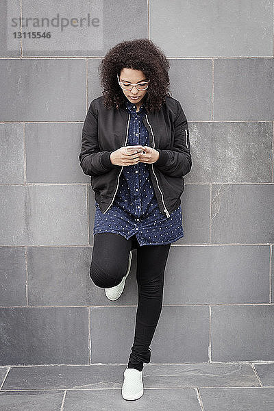 Junge Frau in voller Länge  die ein Smartphone benutzt  während sie sich an eine Betonwand lehnt