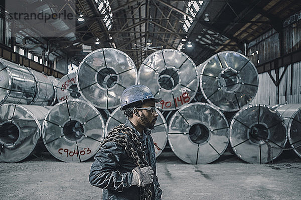 Seitenansicht eines männlichen Arbeiters  der eine Kette trägt  während er an aufgerollten Stahlblechen in der Fabrik steht