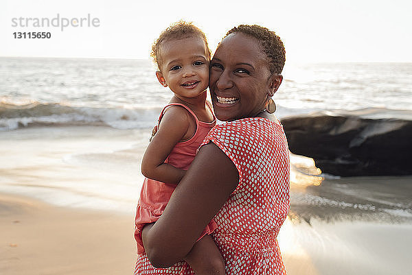 Porträt einer Mutter  die eine Tochter trägt  während sie bei Sonnenuntergang am Strand steht