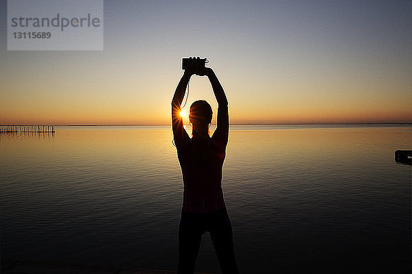 Rückansicht der Silhouette einer Frau  die ein Smartphone in der Hand hält und gegen das Meer trainiert