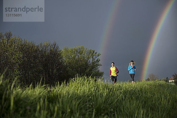 Freunde laufen auf Grasfeld gegen den Himmel mit doppeltem Regenbogen