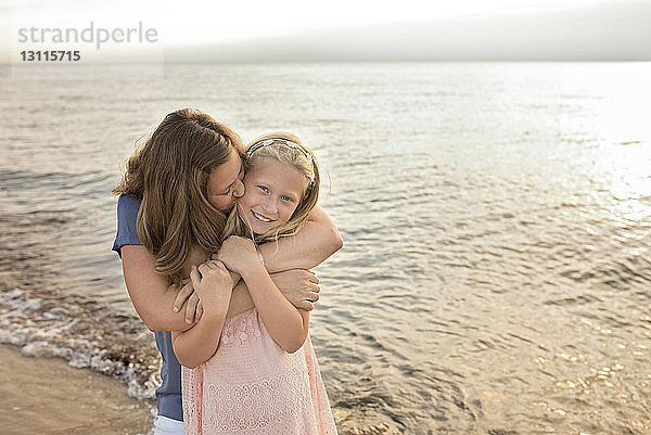 Porträt einer fröhlichen Tochter mit einer Mutter  die sie bei Sonnenuntergang am Strand küsst und umarmt