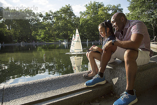 Glücklicher Vater und Tochter spielen mit einem ferngesteuerten Segelboot auf einem Teich im Park