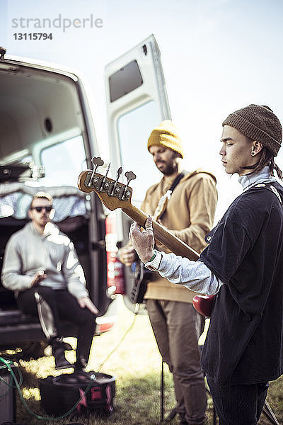 Freunde spielen Gitarre  während ein Mann im Wohnwagen zu Hause auf einem Feld gegen den Himmel sitzt