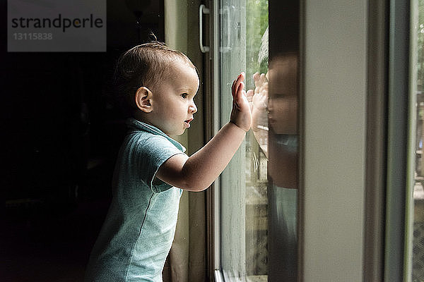 Seitenansicht eines süßen kleinen Jungen  der durch ein Fenster schaut  während er zu Hause in der Dunkelkammer steht