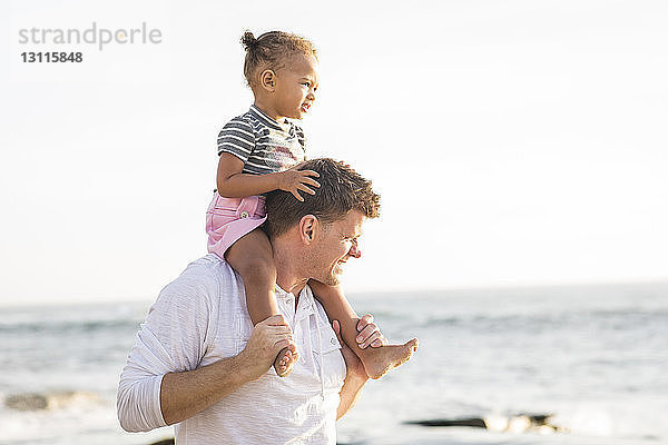 Vater trägt Tochter auf den Schultern  während er am Strand vor klarem Himmel steht