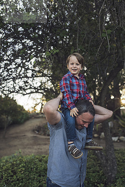 Porträt eines lächelnden Sohnes  der beim Spielen im Park auf den Schultern des Vaters sitzt