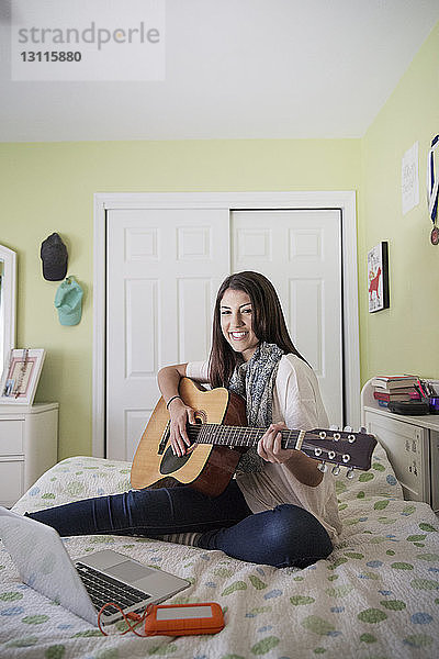 Porträt einer glücklichen Teenagerin  die Gitarre spielt  während sie zu Hause am Laptop sitzt