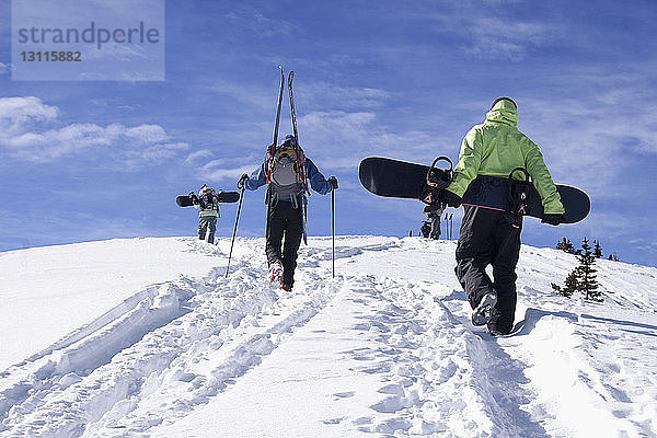 Rückansicht von Freunden  die Snowboards und Skier tragen  während sie auf einem schneebedeckten Berg gehen