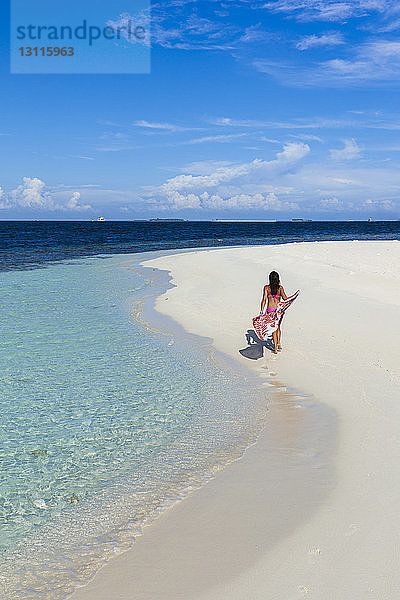 Rückansicht einer Frau mit Sarong  die am Strand vor blauem Himmel am sonnigen Tag spazieren geht