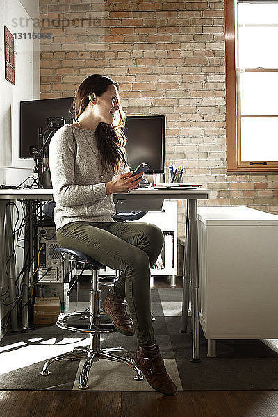 Nachdenkliche Geschäftsfrau schaut weg  während sie im Kreativbüro ein Smartphone in der Hand hält