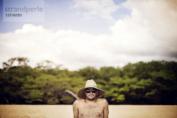 Porträt eines Mannes mit Strohhut  der sich am Strand vor bewölktem Himmel entspannt