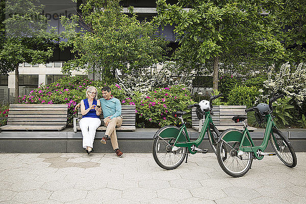 Älteres Paar  das ein Smartphone benutzt und auf einer Bank am Fußweg im Park sitzt