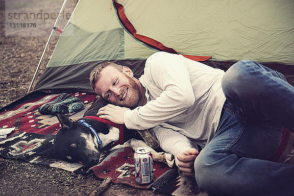 Glücklicher Mann liegt auf Hund vor dem Zelt auf dem Campingplatz