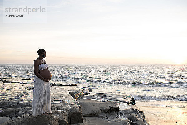 Schwangere Frau  die bei Sonnenuntergang am Strand gegen den Himmel steht