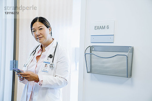 Porträt einer selbstbewussten Ärztin  die ein Smartphone benutzt  während sie im Krankenhaus gegen die Tür steht