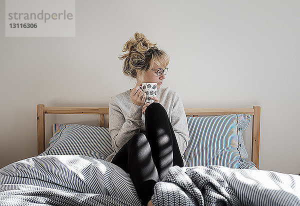 Frau hält Kaffeetasse  während sie zu Hause auf dem Bett sitzt