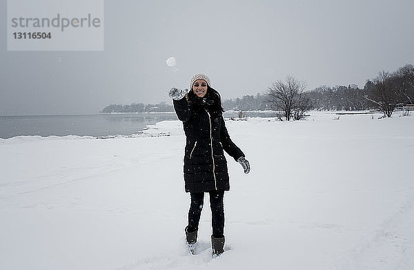 Porträt einer lächelnden Frau  die einen Schneeball wirft  während sie am schneebedeckten Strand gegen den Himmel steht