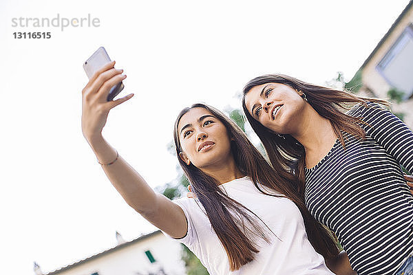 Niedriger Blickwinkel auf Freunde  die sich mit dem Mobiltelefon selbstständig machen  während sie gegen den klaren Himmel stehen