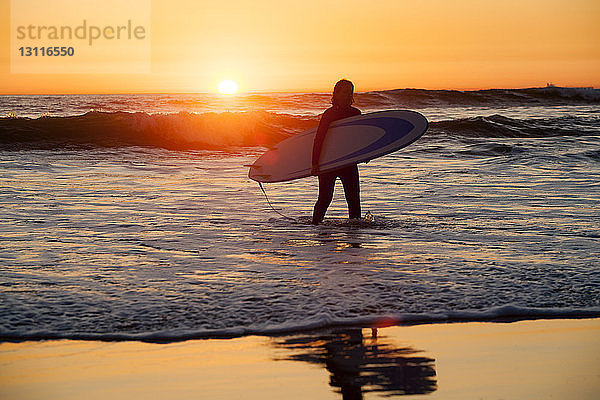 Frau trägt Surfbrett  während sie bei Sonnenuntergang am Ufer spazieren geht