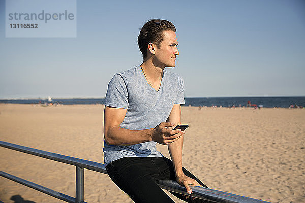 Junger Mann sitzt am Strand auf einem Geländer vor klarem Himmel