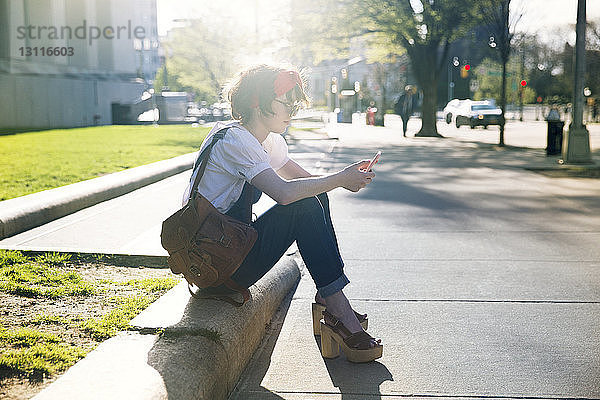 Seitenansicht einer Frau  die ein Smartphone benutzt  während sie an einem sonnigen Tag auf einem Fußweg sitzt