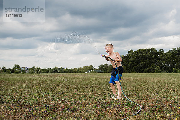 Verspielter Junge in voller Länge ohne Hemd  der mit einem Gartenschlauch auf einem Grasfeld vor bewölktem Himmel im Hinterhof spielt