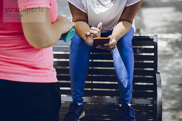 Niedriger Teil einer Frau  die ein Smartphone benutzt  während sie auf einer Bank sitzt und ihr Freund im Vordergrund steht