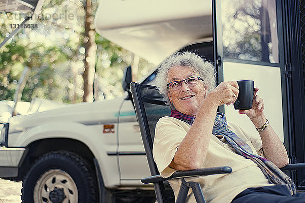 Glückliche ältere Frau hält Kaffeetasse in der Hand  während sie in einem Wohnwagen im Wald sitzt
