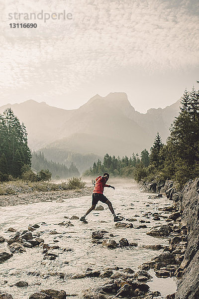 Wanderer springt in voller Länge auf Felsen  während er den Fluss gegen die Berge überquert