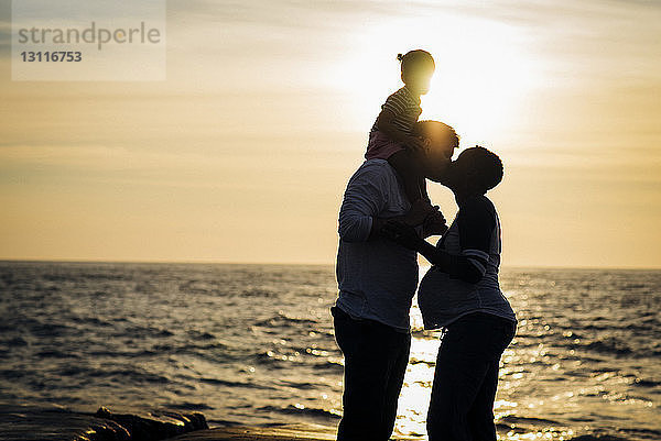 Scherenschnitt-Mann küsst seine Frau  während er seine Tochter bei Sonnenuntergang am Strand auf den Schultern trägt