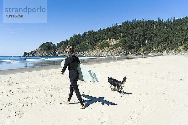 Mann hält Surfbrett mit Hund beim Spaziergang am Strand