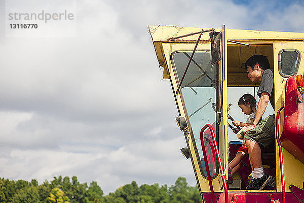 Seitenansicht von Vater und Tochter im Traktor auf dem Bauernhof vor bewölktem Himmel