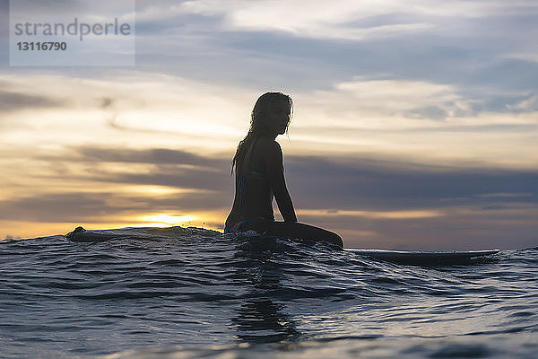 Seitenansicht einer jungen Frau  die bei Sonnenuntergang auf einem Surfbrett im Meer vor bewölktem Himmel sitzt